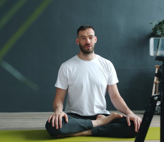 “거실이 곧 개인 훈련장”… AI 카메라와 홈트의 결합, ‘공간의 해방’ Man meditating on yoga mat recording with phone.